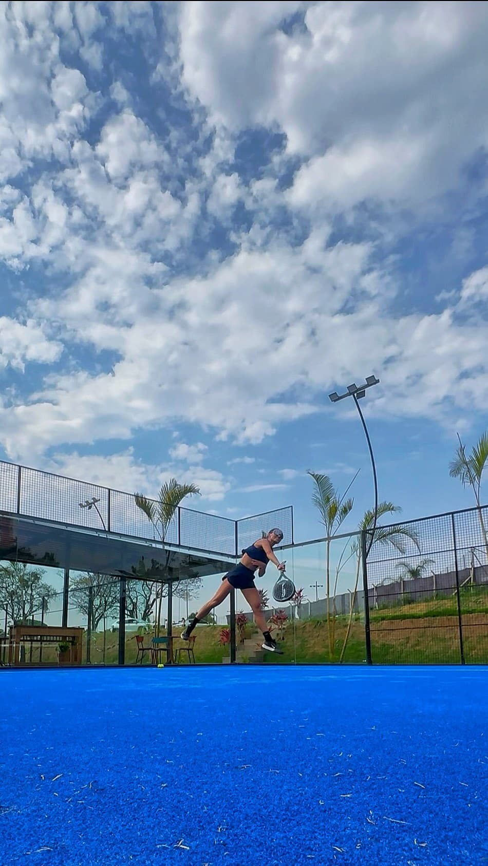 Jogadora sacando na quadra de padel azul da Arena M33, vista de baixo com o céu cheio de nuvens