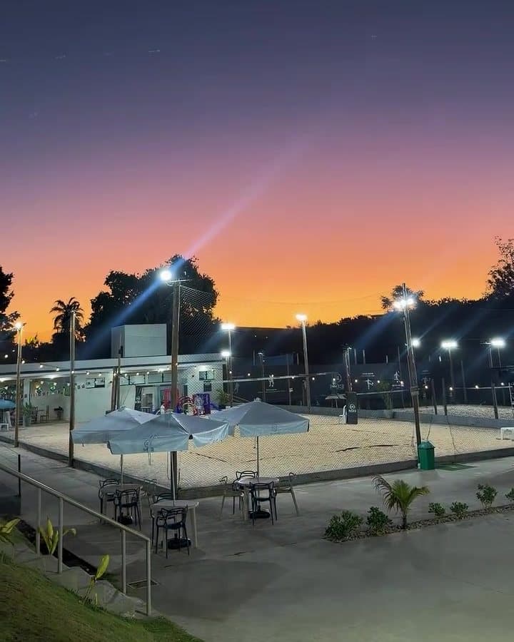 Mesas com guarda-sol ao lado das quadras de areia da Arena M33, no fim da tarde, com os refletores acesos e o céu laranja
